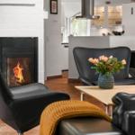 Wohnbereich mit Kamin, schwarzen Sesseln und Holztisch mit Blumen.