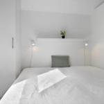 Schlafzimmer mit weißem Bett, grauem Kissen und weißen Schränken. Auf Regal eine Pflanze und zwei Wandlampen.
