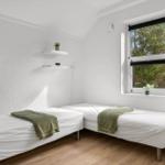 Zwei Einzelbetten mit grünen Decken, Fenster mit Blick auf Bäume und weiße Wände.