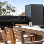 Holztisch mit Stühlen auf Terrasse. Hintergrund: moderne Hauswand und Baum.