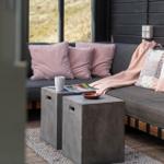 Ein Sofa mit Kissen und Decke vor großen Fenstern. Zwei Betonbeine stehen daneben.
