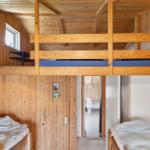 Zimmer mit Doppelstockbett und zwei Einzelbetten. Holzwände und Türöffnung zum Bad.