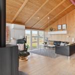 Wohnzimmer mit Holzdecke, Sofa, Sessel und Kaminofen. Große Fenster mit Blick auf die Landschaft.