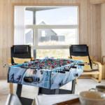 Ein Foosballtisch steht im Wohnzimmer mit Holzwänden und großen Fenstern.