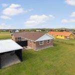 Ein Haus mit Carport auf grünem Grundstück, umgeben von weiteren Häusern und Dünen.
