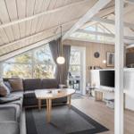 Wohnbereich mit Sofa, Tisch und TV. Küche mit Arbeitsplatte. Holzdecke und große Fenster.
