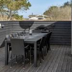 Terrasse mit Tisch, Stühlen und Grill. Hintergrund: Holzwand und Bäume.