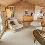 Wohnzimmer mit Holzwand, Sessel, Couchtisch und Küchenbereich im Hintergrund.