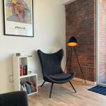 A dark chair stands before a brick wall. Beside it is a floor lamp and a bookcase.