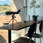 A table with binoculars, chair, and plant by window with view of landscape.