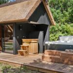 Holzterrasse mit Whirlpool und Holzbadewanne vor einem strohgedeckten Haus.