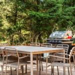 Terrasse mit Tisch, Stühlen und Grill. Hintergrund: grüne Bäume und Pflanzen.