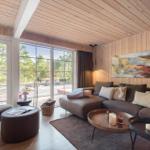 Wohnzimmer mit Holzwand, großem Sofa und Blick auf Balkon und Wald.