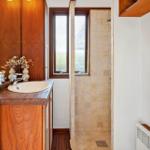 Bad mit Dusche, Waschbecken und Holzmöbeln. Zwei Fenster sorgen für Tageslicht.