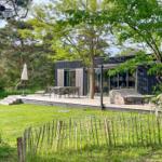 Moderne Villa mit Terrasse, Garten und Bäumen. Außentisch, Sonnenschirm und Grilldecke sichtbar.