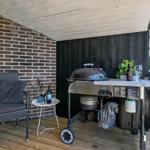 Terrasse mit Grill, Stühlen und Tisch. Holzdecke, Ziegelwand und Pflanzen.