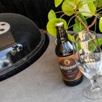 Eine Flasche Bier und ein Glas stehen auf einer Terrasse neben einem Grill und einer Pflanze.