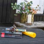 Auf einem Tisch liegen Buch, Sonnenbrille und Sonnenschutzcreme. Im Hintergrund Blumen und Kerze.