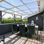 Terrasse mit Tisch und Stühlen unter Glasdach. Holzdecke, dunkle Wand und grüner Hintergrund.
