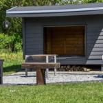 Ein graues Holzhaus mit offener Tür und Brennholz darunter. Draußen steht ein Tisch und Bänke.