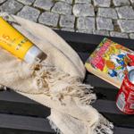 Sonnencreme, Getränk und Comic auf einer Bank vor Steinplatten.