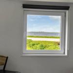 Durch das Fenster sieht man einen See mit Grünflächen und einem Weg. Zwei Stühle stehen im Raum.
