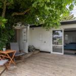Terrasse mit Tisch und Stühlen unter einem großen Baum. Eingang und Schiebetür führen ins Haus.