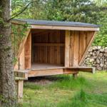 Holzkonstruktion mit Dach, umgeben von Bäumen und einem Holzstapel.