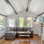 Ein Wohnzimmer mit grauem Sofa, Holzboden und weißen Wänden. Große Fenster mit Blick auf Grün.