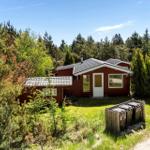 Rotes Holzhaus mit weißen Fensterrahmen und Dach im Wald. Grasfläche und Weg führen zum Eingang.