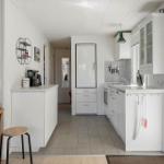 Küche mit weißen Schränken, Kühlschrank und Arbeitsplatte. Links Wohnbereich mit Sofa und Stuhl.