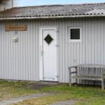 House with gray siding, white door, and wooden bench in front. Sign with name and number.