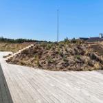 Holzterrasse mit Steigstufen zu einer Dünenlandschaft mit Blick auf Häuser und einen Mast.