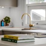 Kücheninsel mit Kochbuch und Gläsern. Hintergrund: goldener Wasserhahn und Fenster.
