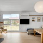 Wohnzimmer mit Holzfußboden, Fernseher, Sitzbank und Schaukelstuhl. Große Fenster und Deckenlampe.