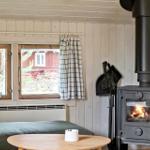Room with wood stove, window with curtains and table. Outside, a red building is visible.
