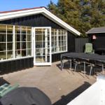 Haus mit Terrasse, Tisch, Stühlen und Gartenmöbeln. Schwarze Fassade, weiße Fenster und roter Dachrand.