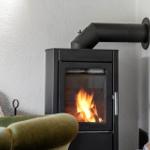 A wood-burning stove with burning logs, next to a green armchair and a stack of firewood.