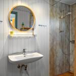 Bathroom with sink, round mirror, and shower with wooden shelf.