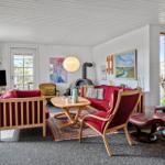 Living room with red sofa, wooden furniture, and fireplace. White walls, large windows, and wall art.