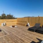 Holzterrasse mit Liegen, Sitzbank und Feuerkorb. Umzäunt mit Holz und Blick auf Bäume.