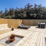 Holzterrasse mit Liegen, Esstisch und integriertem Feuerkorb. Hintergrund: Holzzaun und Nadelbäume.