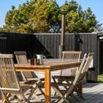 Holztisch mit Stühlen auf Terrasse, Grill und Hinterhof mit Bäumen.