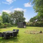 Grüner Garten mit Tisch und Stühlen, kleiner Holzschuppen und Bäumen im Hintergrund.