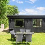 Schwarzes Holzhaus mit Gartenmöbeln und Grill. Grün umgeben unter blauem Himmel.