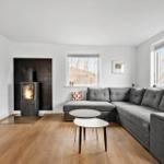 Wohnzimmer mit grauem Sofa, Kamin und Holzboden. Fenster mit Blick auf Bäume.