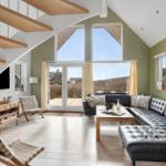Wohnzimmer mit schwarzer Ledercouch, Holztisch und großen Fenstern. Holzstufen führen zur oberen Etage.