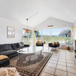 Wohnzimmer mit Sofa, Tisch und Sesseln. Große Fensterfront mit Blick auf Landschaft.