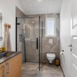Badezimmer mit Dusche, Waschbecken und WC. Graue Fliesen, Spiegel mit Beleuchtung.