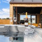 Hochwertiger Whirlpool mit Champagner und Gläsern auf einer Terrasse vor einem Holzhaus.